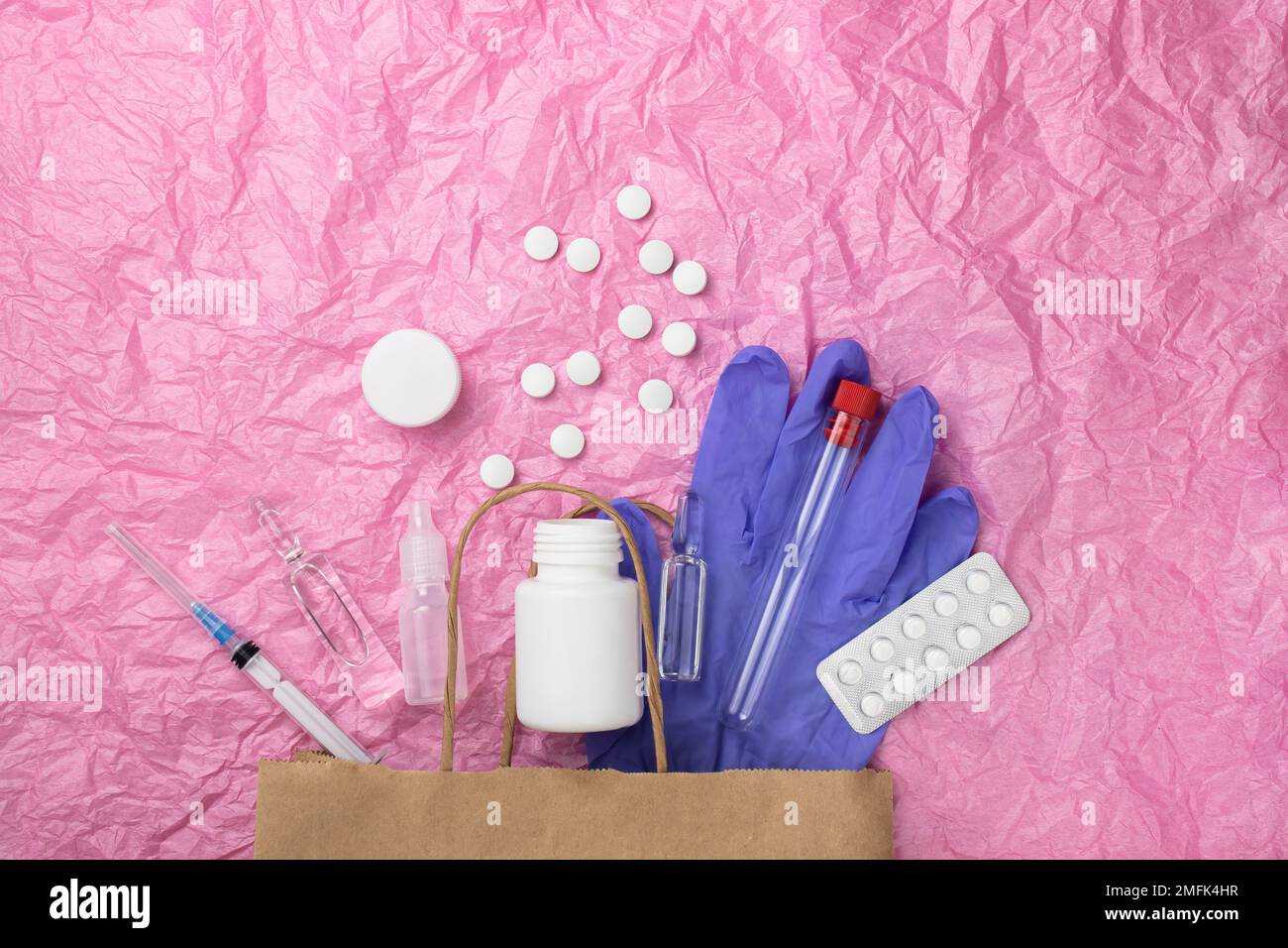 Articoli medici come le fiale della pillola, una siringa e i guanti sbirciano fuori da un sacchetto di carta contro uno sfondo di carta rosa stropicciata. Un sacco di spazio vuoto fo Foto Stock