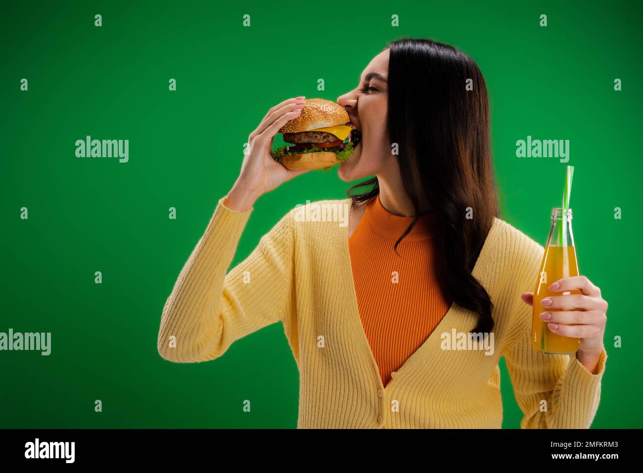 donna brunette tenere una bottiglia di limonata fresca e mangiare gustoso hamburger isolato sul verde, immagine stock Foto Stock