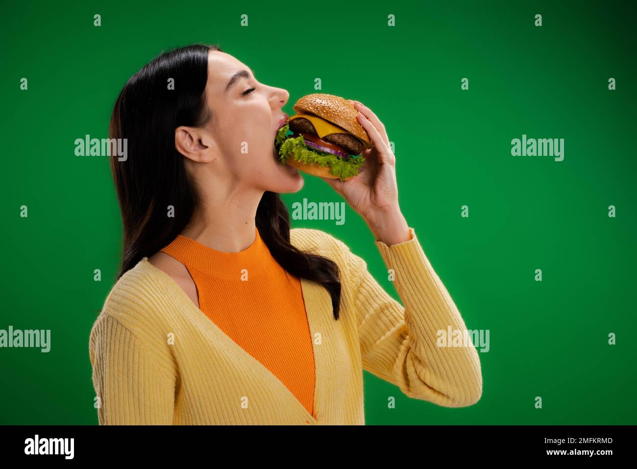 bruna donna affamata mangiare delizioso hamburger con carne e formaggio isolato sul verde, immagine stock Foto Stock
