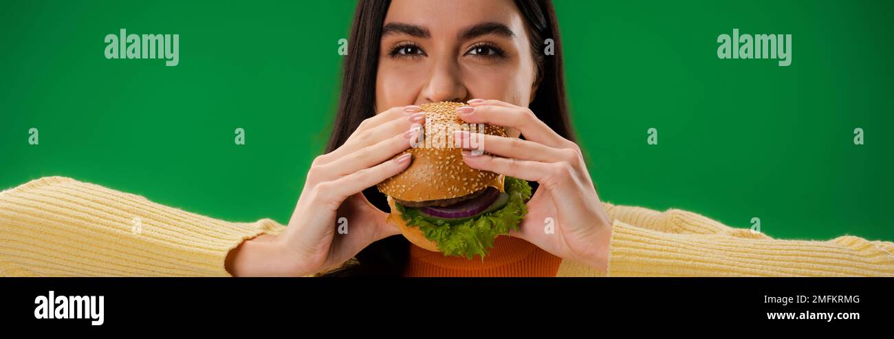 giovane e donna affamata mangiare hamburger e guardare la macchina fotografica isolato su verde, banner, immagine stock Foto Stock