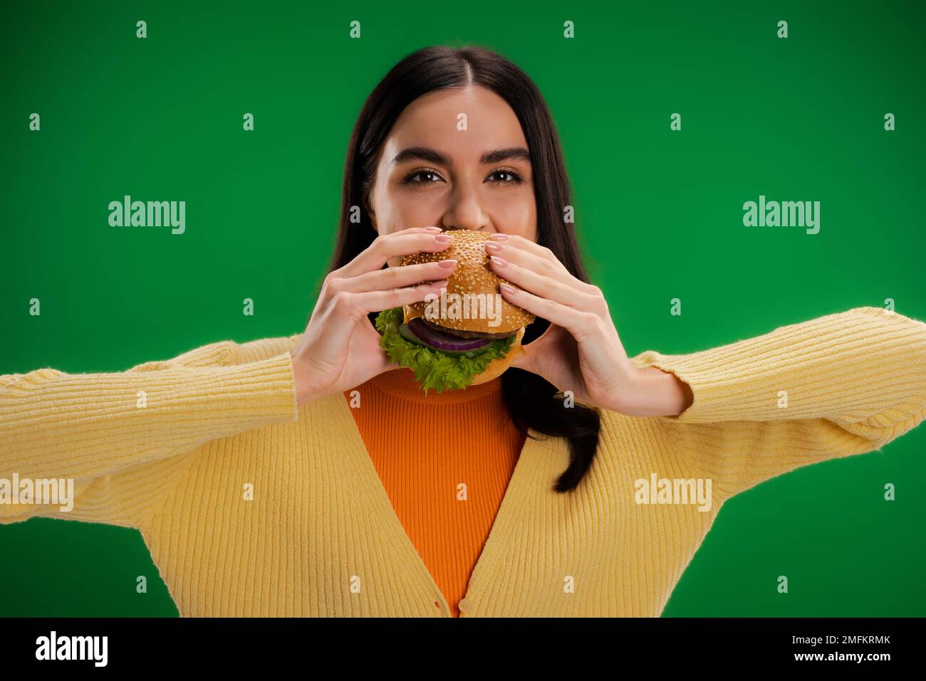 giovane donna brunette mangiare delizioso hamburger e guardare la macchina fotografica isolato sul verde, immagine stock Foto Stock