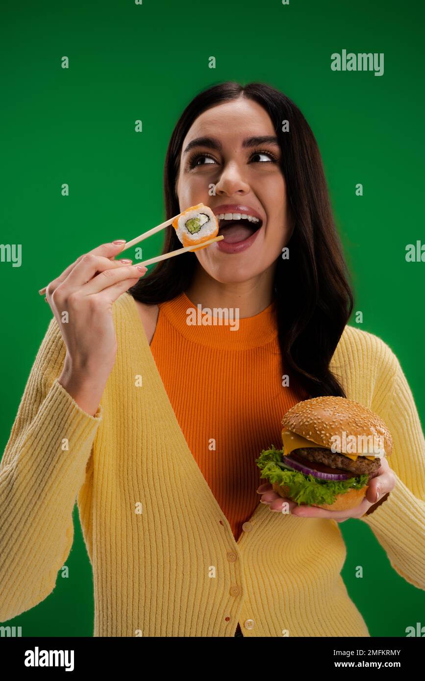 donna soddisfatta che tiene hamburger e mangiare sushi fresco roll con bacchette isolato sul verde, immagine stock Foto Stock