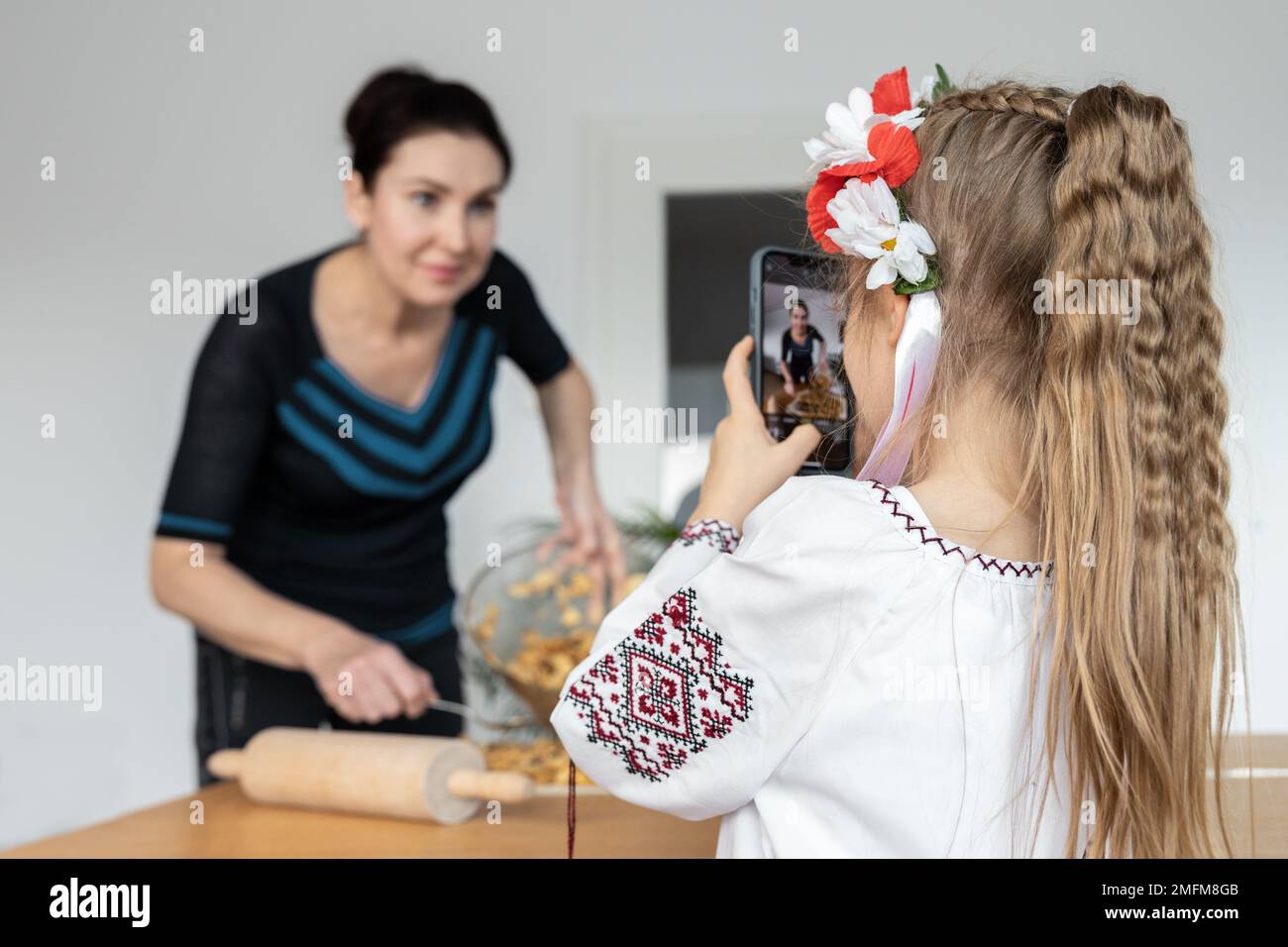 Una ragazza con capelli castani in costume ucraino fotografa sua madre, che sta cucinando torta di mele sul tavolo Foto Stock