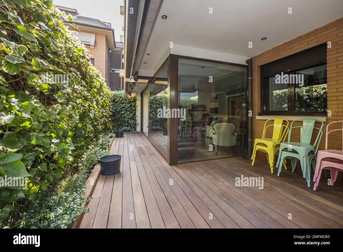 Terrazza di una casa al piano terra con siepi e piante decorative, pavimenti in acacia, divisori in vetro e sedie colorate in metallo Foto Stock