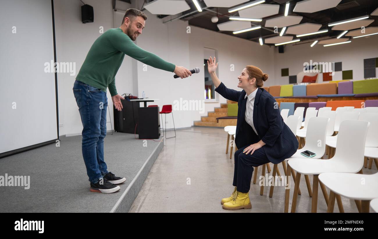 Una donna d'affari caucasica dai capelli rossi siede in prima fila in una sala conferenze vuota. L'uomo barbuto fa una domanda nella sala. Foto Stock