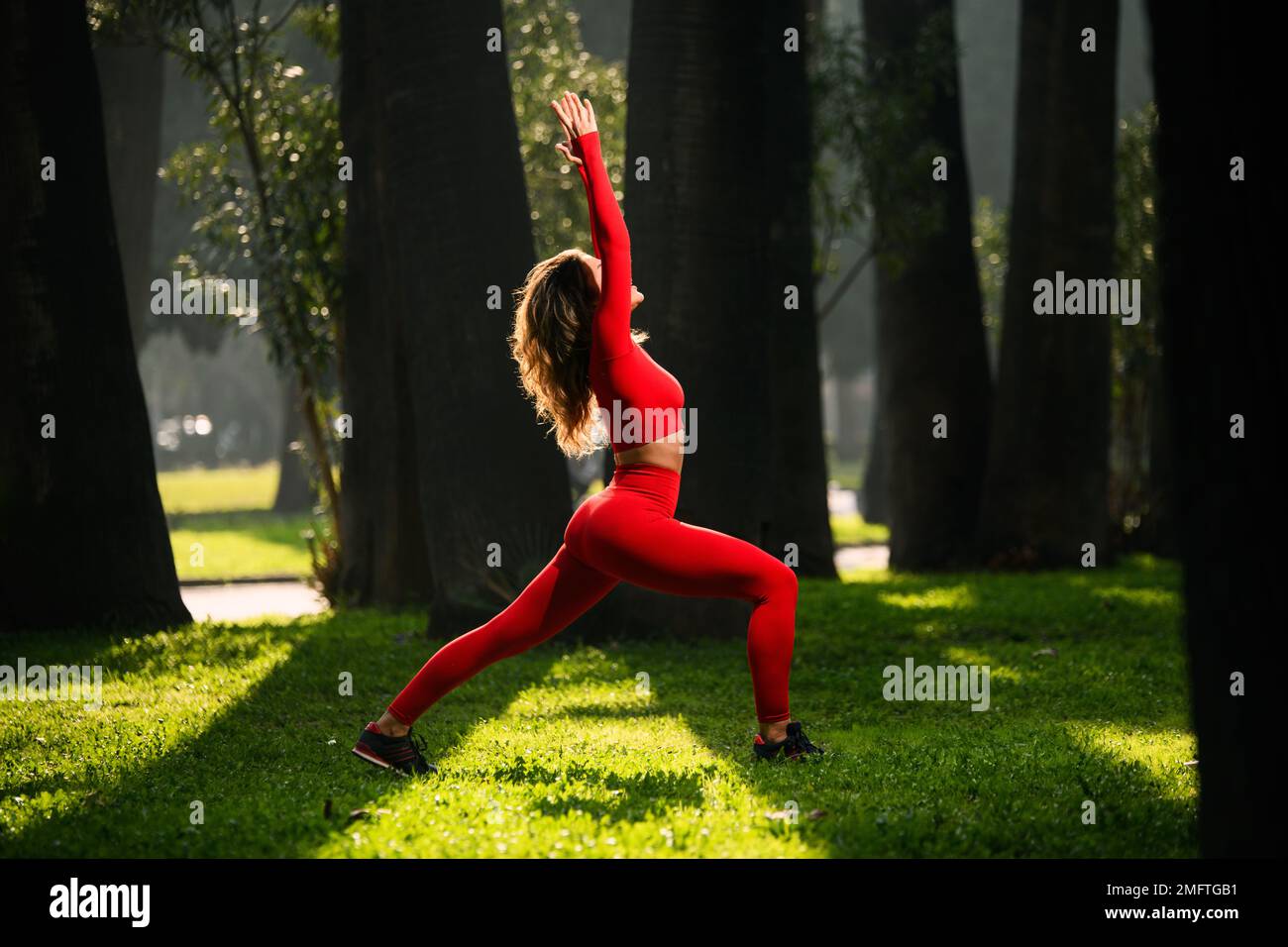 Donna con abito da yoga di colore rosso, che posa i movimenti del guerriero di yoga su uno sfondo verde grasss Foto Stock