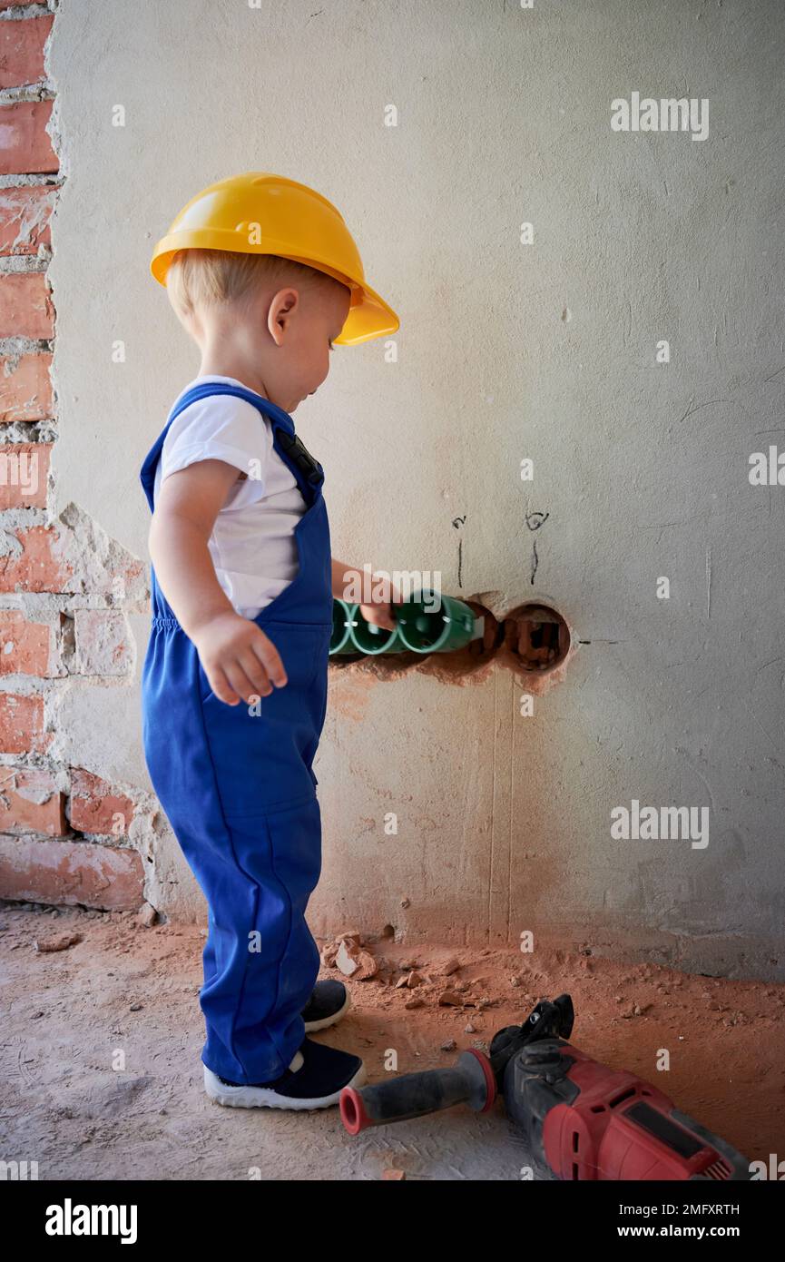 Piccolo elettricista che installa canaline per cavi e prese a muro. Bambino in tute di lavoro e di sicurezza costruzione casco montaggio cablaggio elettrico in appartamento in fase di ristrutturazione. Foto Stock