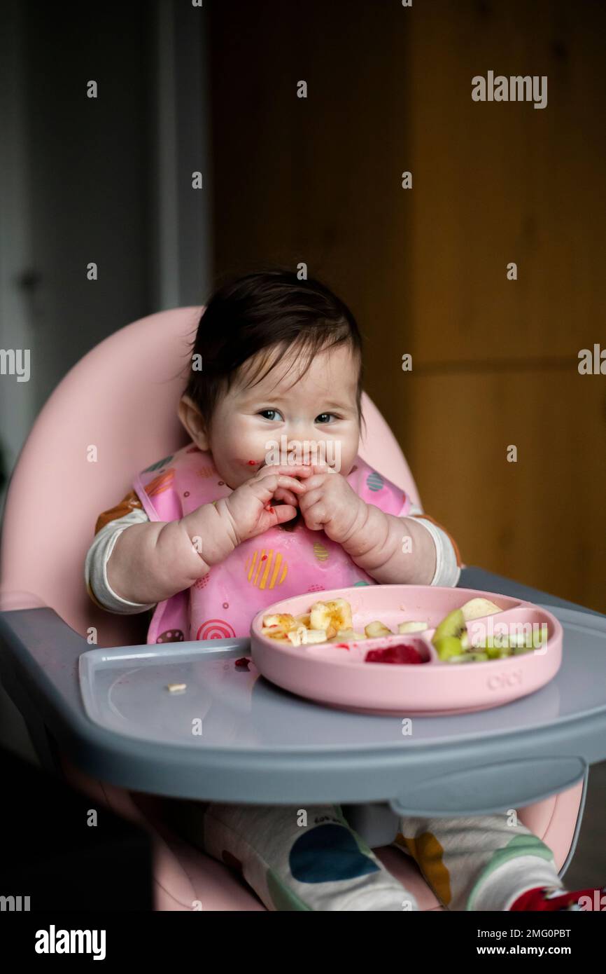 Ragazza cute del bambino che siede sul highchair in cucina, mangiando le verdure grezze, la frutta e sorridente. Mangiare sano per il concetto del bambino. Stile di vita quotidiano della mamma. Foto Stock