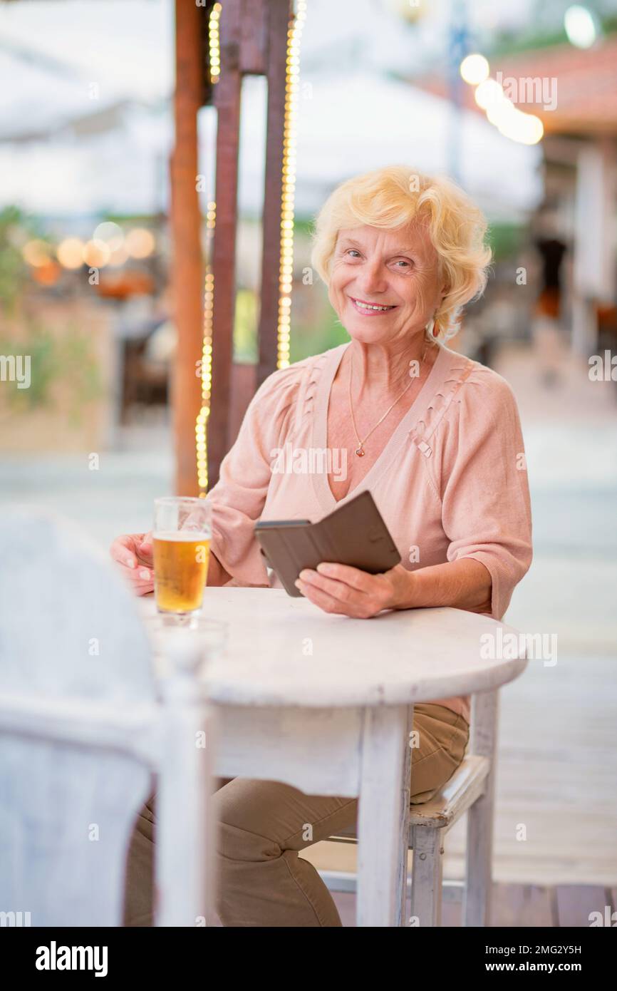 Donna matura seduta a un tavolo in un bar estivo e bere birra Foto Stock