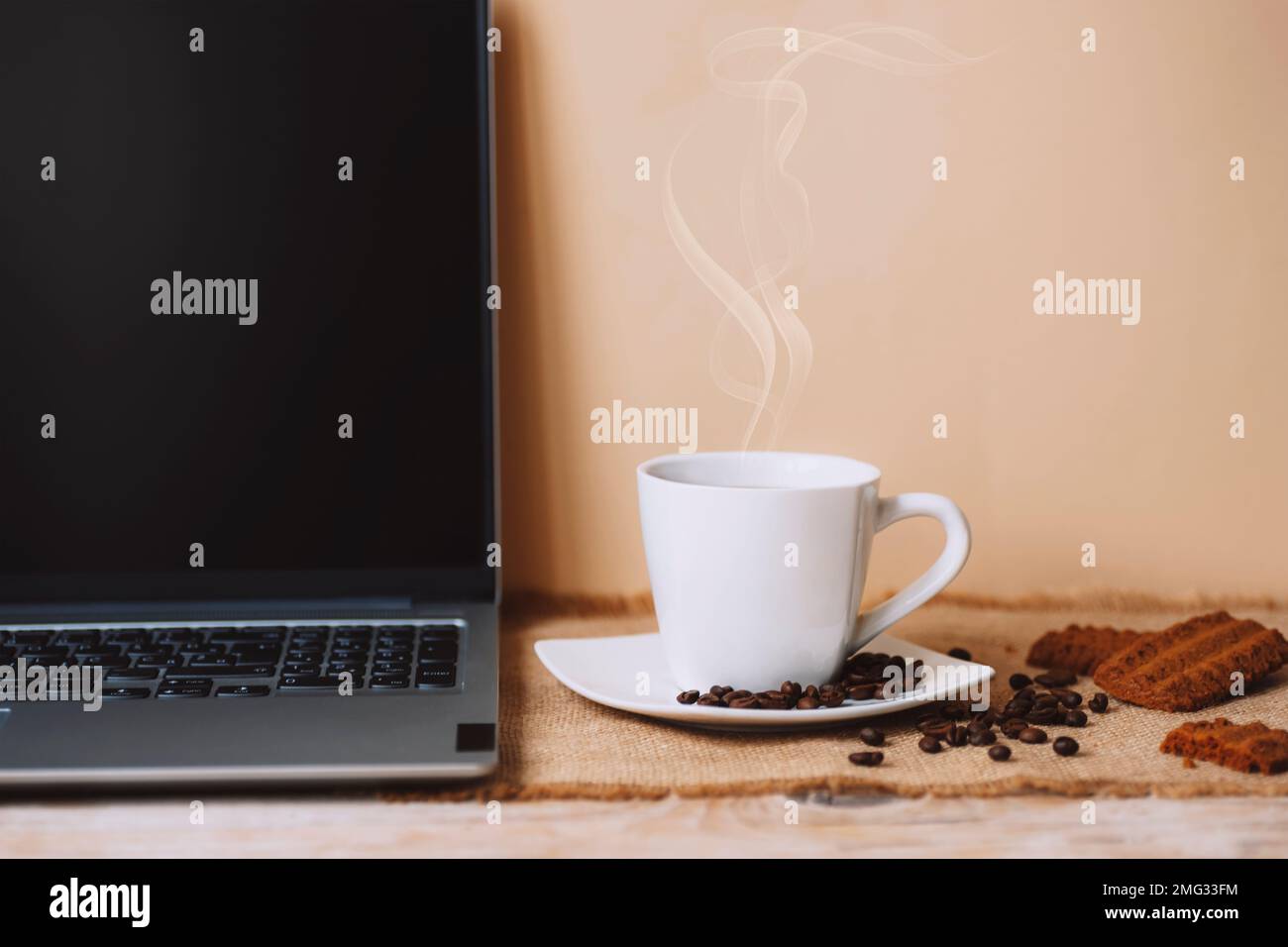 Tazza bianca di caffè caldo con chicchi, biscotti e computer portatile su sfondo da tavolo beige. Foto Stock