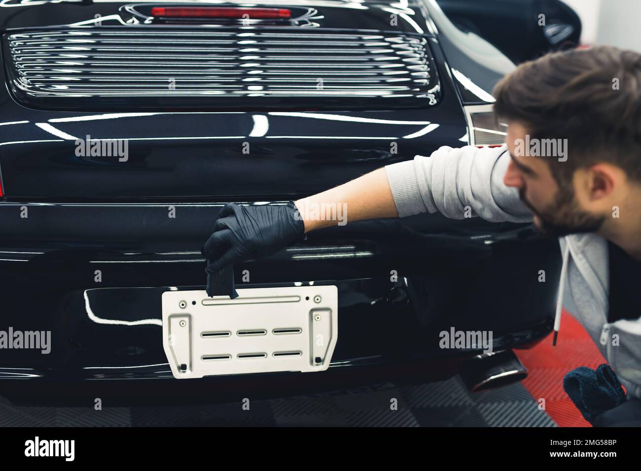 Uomo bianco che indossa guanti neri che squattano applicando il rivestimento in ceramica sul retro dell'auto nera. Dettagli di auto professionali. Ripresa orizzontale da interno . Foto di alta qualità Foto Stock