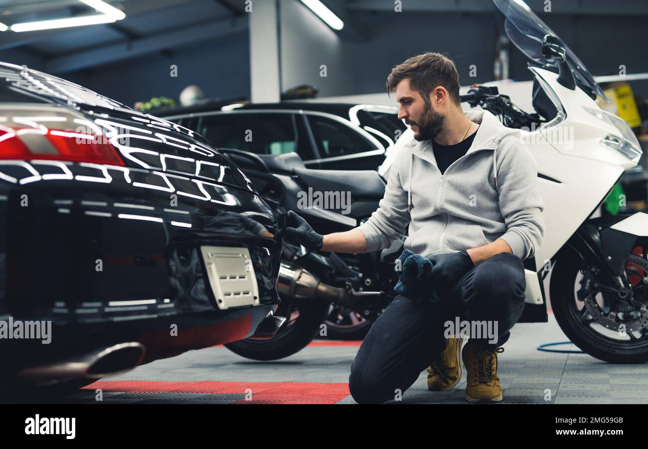 Uomo bianco che si inginocchia applicando il rivestimento in ceramica alla parte posteriore di un'auto nera con la motocicletta sullo sfondo. Dettagli di auto professionali. Garage. Scatto orizzontale in interni. Foto di alta qualità Foto Stock