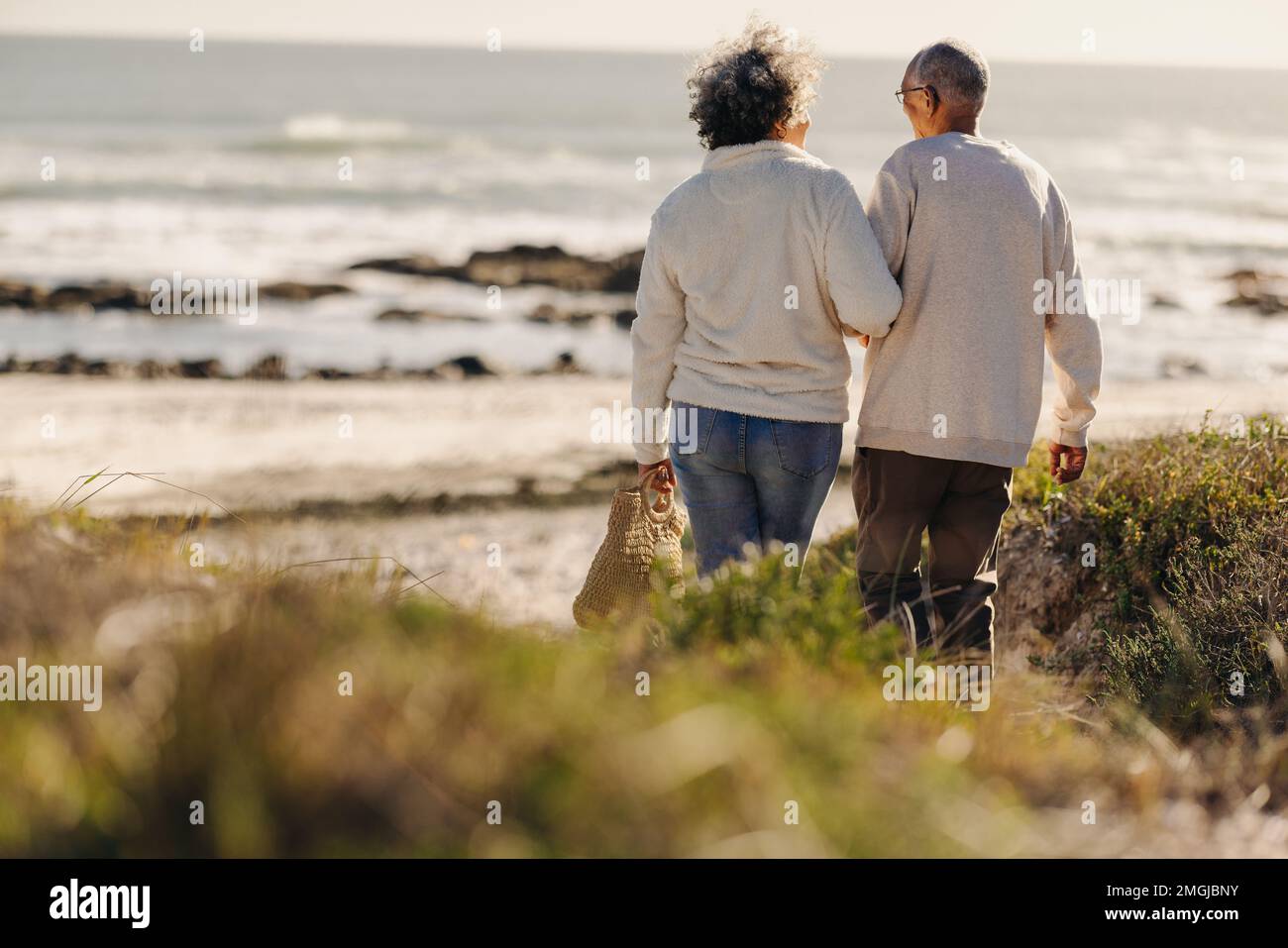 Vista posteriore di una romantica coppia anziana che va verso la spiaggia per un picnic. Felice coppia anziana che prende una vacanza al mare dopo il pensionamento. Foto Stock