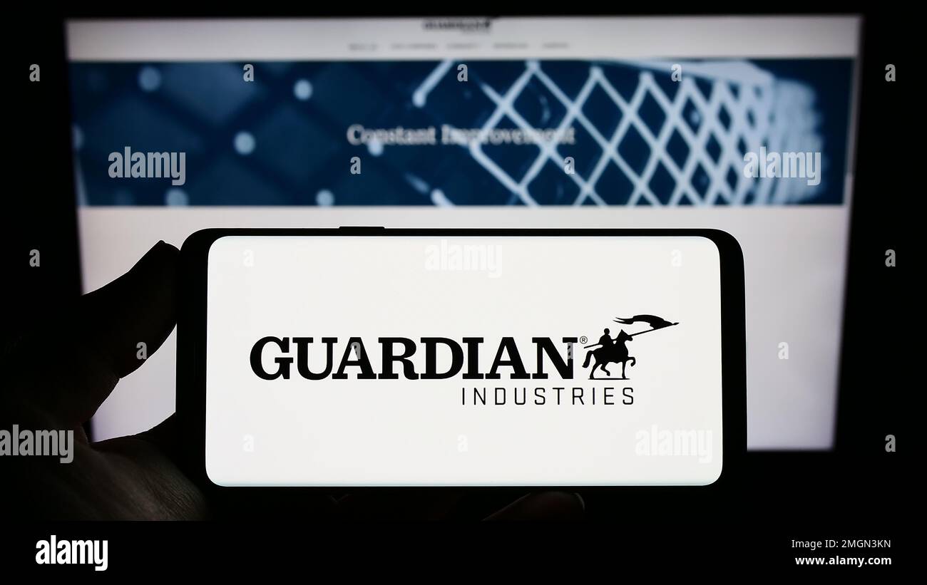 Persona che tiene in mano uno smartphone con il logo dell'azienda produttrice di vetro statunitense Guardian Industries sullo schermo di fronte al sito Web. Messa a fuoco sul display del telefono. Foto Stock