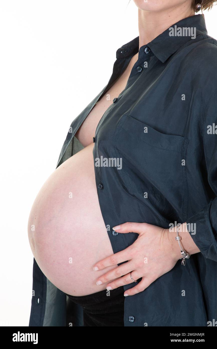 Donna incinta in camicia tiene le mani sul ventre su uno sfondo bianco nel concetto di aspettativa di gravidanza Foto Stock