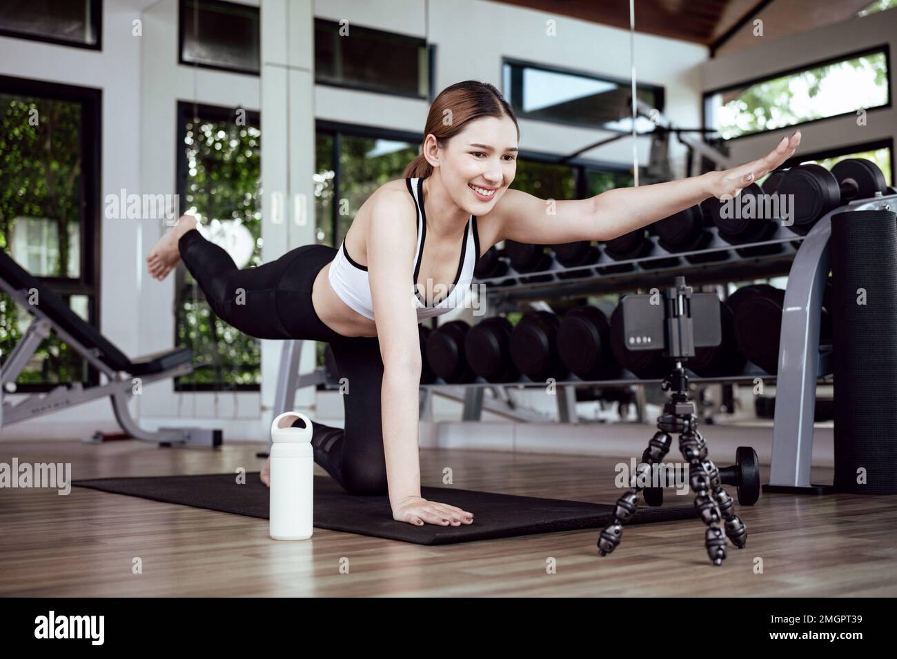 Allenamento femminile asiatico sul materassino yoga e con il telefono cellulare. Blogger sportivo femminile per la registrazione di video tramite smartphone. Esercitazione corso di formazione online Foto Stock