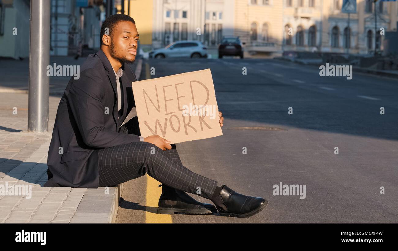 Triste disperato frustrato etnico lavoratore ufficio di un dipendente afro-americano uomo maschio ragazzo seduto su gradini all'aperto tenendo cartoncino cartello poster bisogno di lavoro Foto Stock