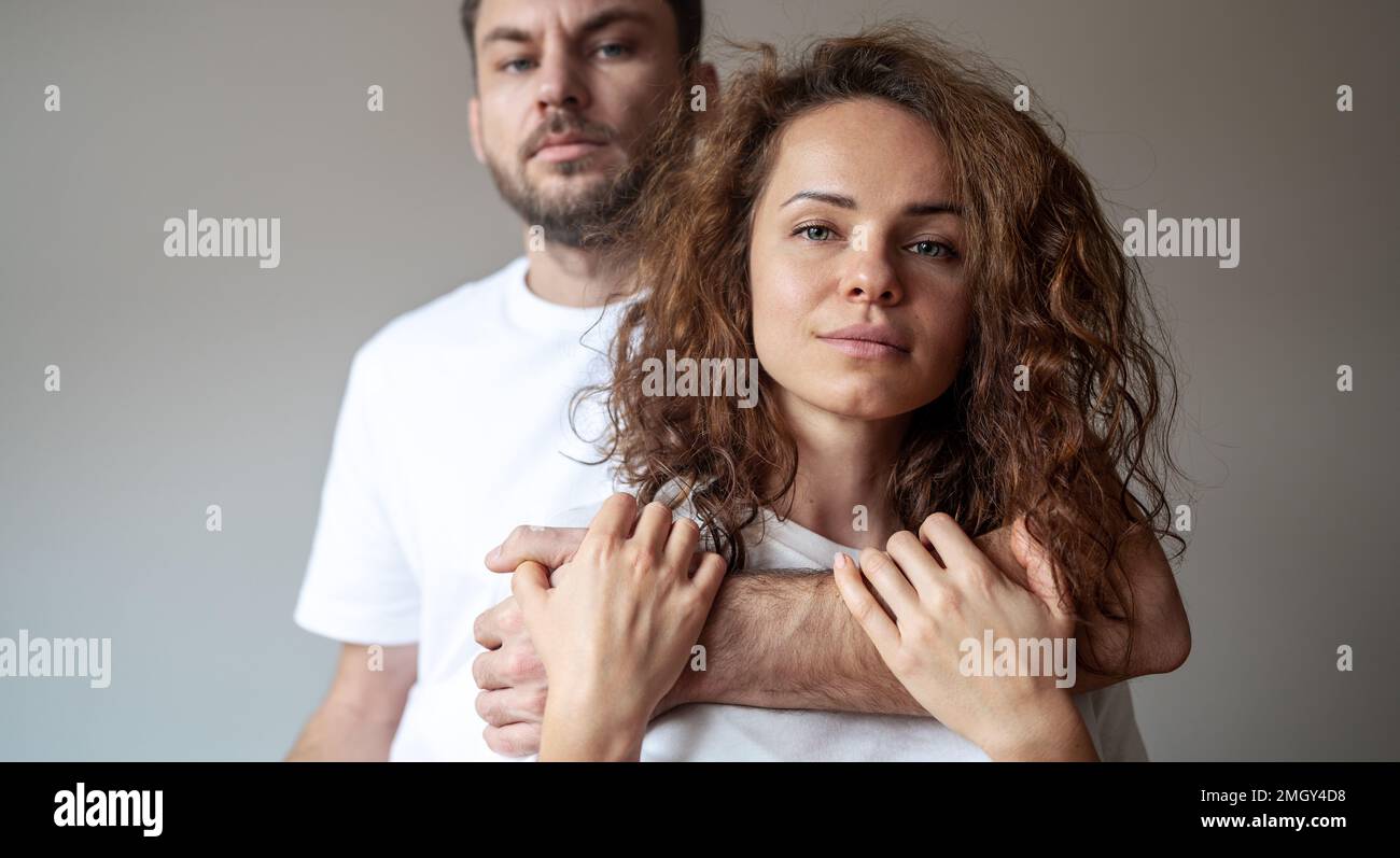 Ritratto donna capelli castani ricci e uomo bearded in piedi dietro e abbracciando donna con la mano, coppia indossando t-shirt bianche guardando la macchina fotografica. Foto Stock