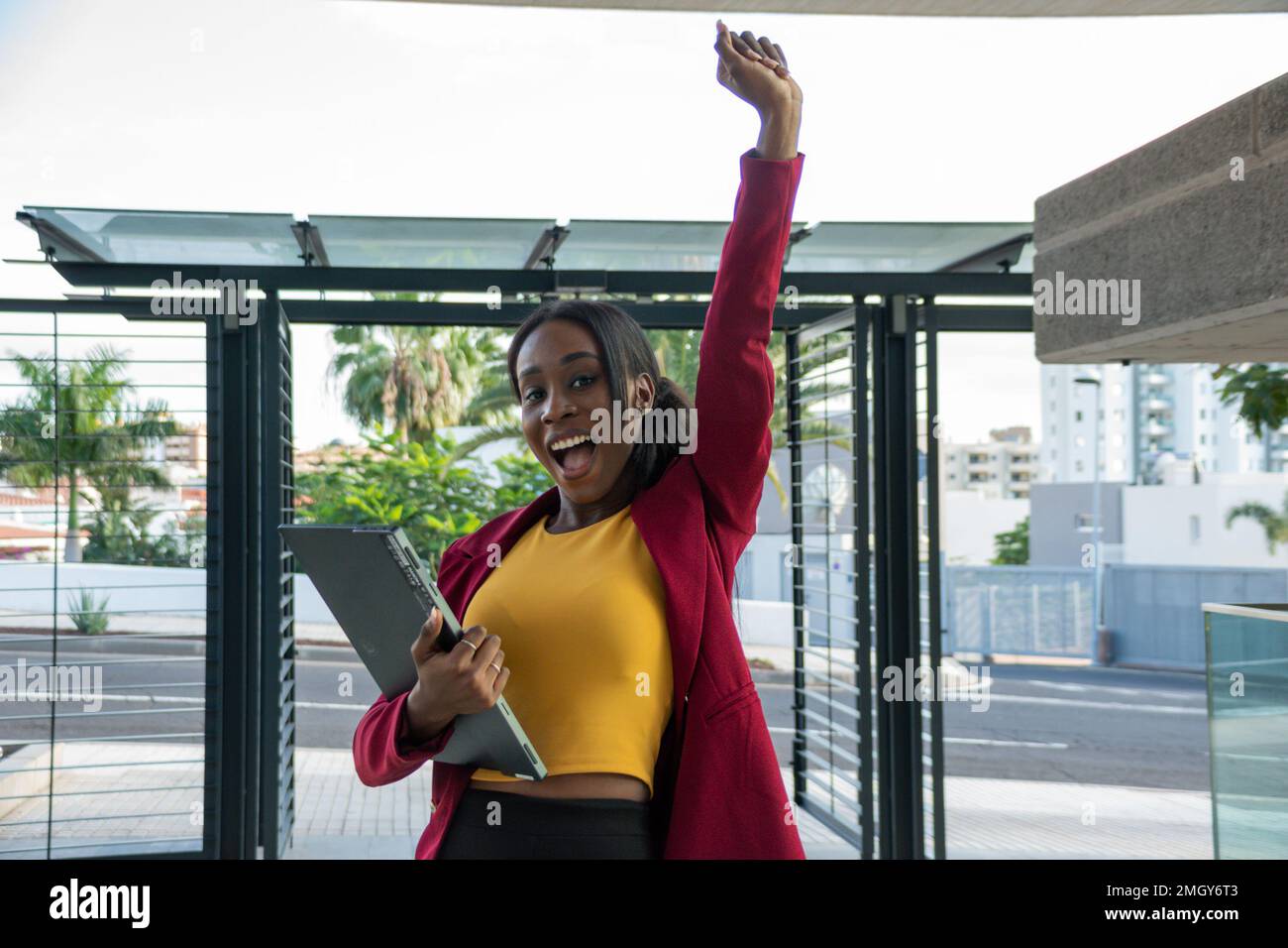 Attraente giovane donna d'affari africana con un braccio sollevato, entusiasta per il successo del lavoro. Tiene la sua tavoletta nell'altra mano Foto Stock