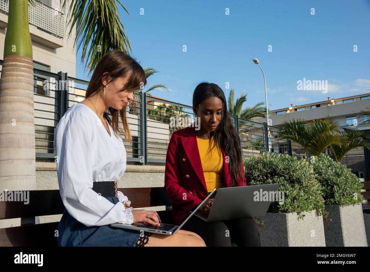 Due giovani donne in viaggio d'affari lavorano all'aperto seduti su una panca a digitare con il loro notebook. Concetto di nomade digitale Foto Stock