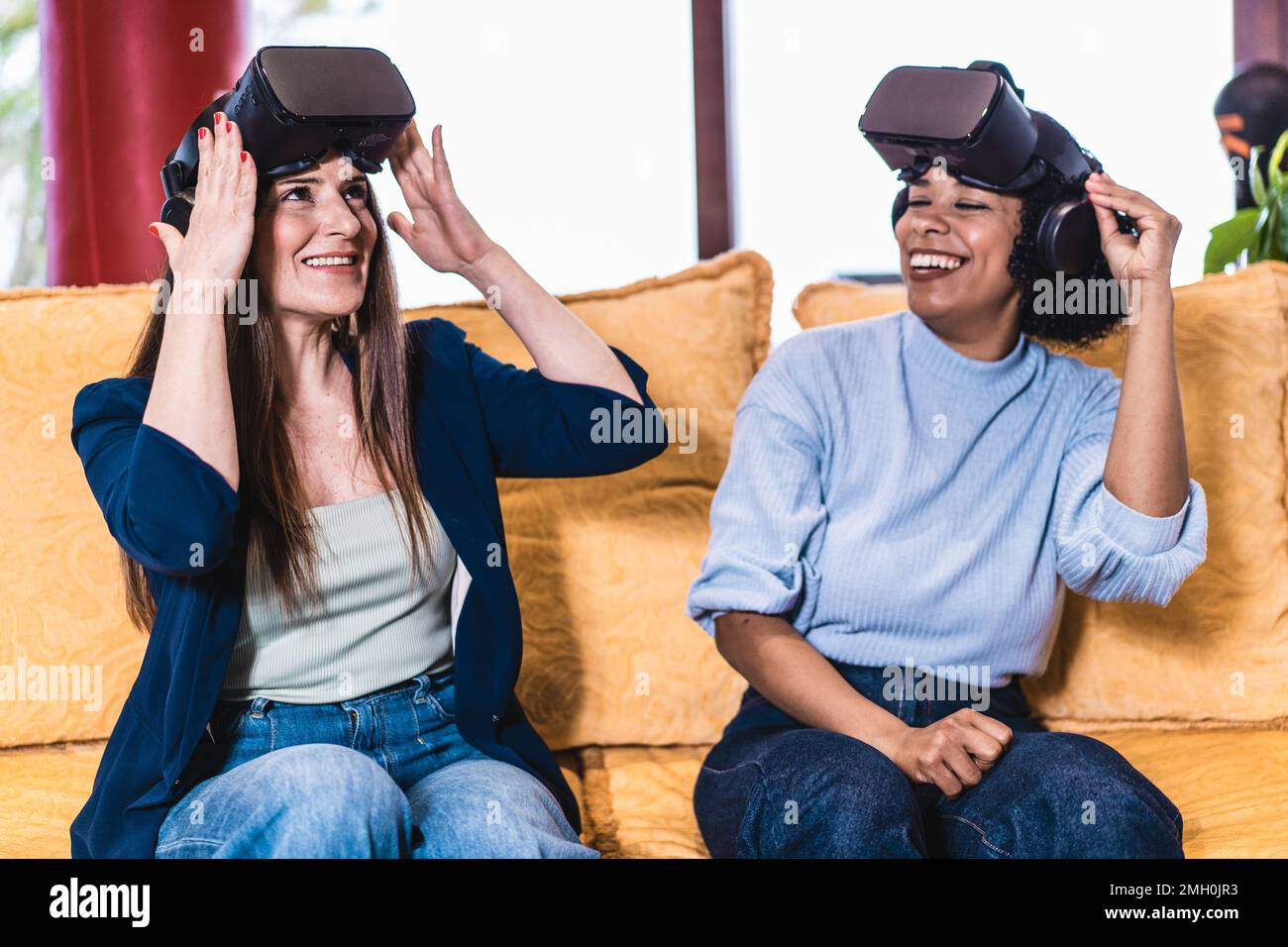 Due amici, uno caucasico e uno brasiliano, si siedono su un divano e hanno appena rimosso i visore VR dalla testa. Stanno ridendo insieme dopo l'expe Foto Stock