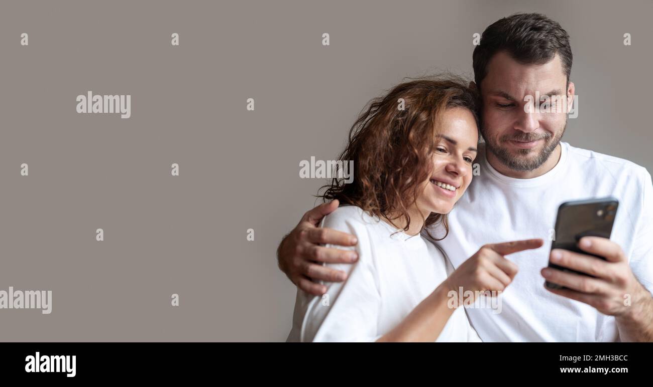 Giovane coppia che indossa T-shirt bianche ricci donna e uomo con beared insieme utilizzando il telefono cellulare e donna sorridente che indica lo schermo del telefono cellulare. Foto Stock
