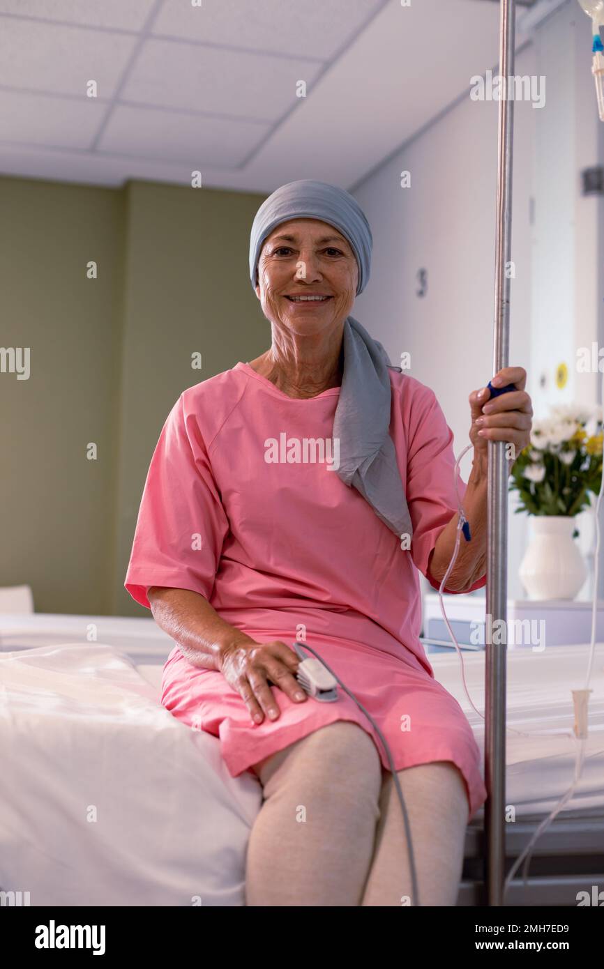 Felice paziente caucasica femminile di cancro con sciarpa della testa seduta sul letto con gocciolamento in ospedale Foto Stock
