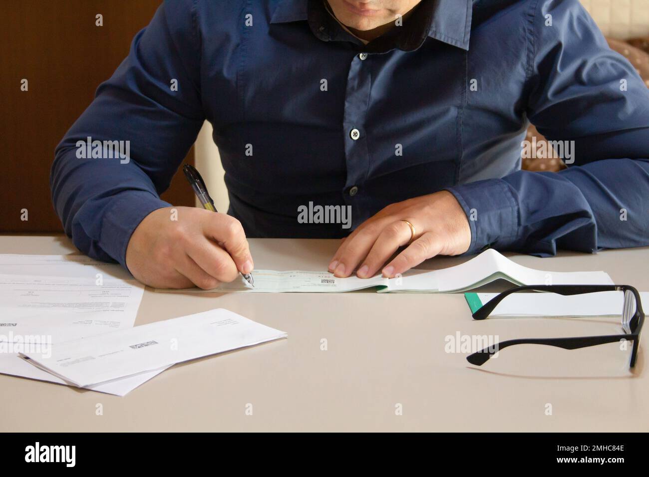 Immagine di un uomo seduto dietro una scrivania con fatture e fatture sparse intorno alla firma di un assegno di pagamento. Foto Stock