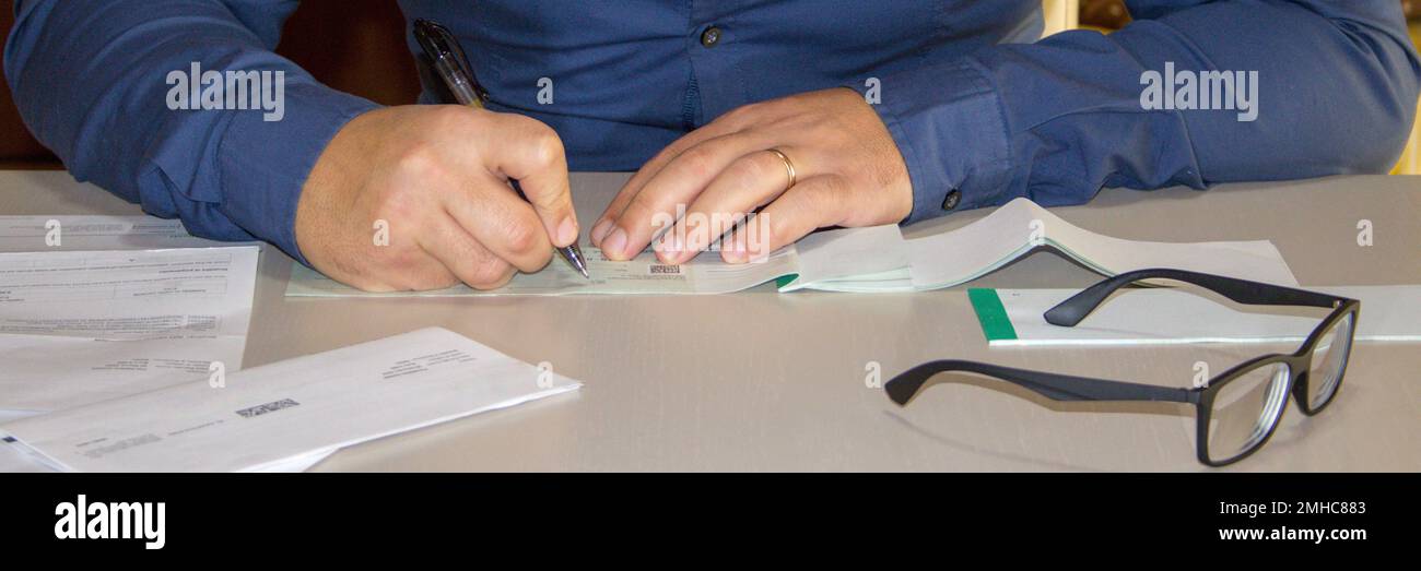 Immagine di un uomo seduto dietro una scrivania con fatture e fatture sparse intorno alla firma di un assegno di pagamento. Banner orizzontale Foto Stock