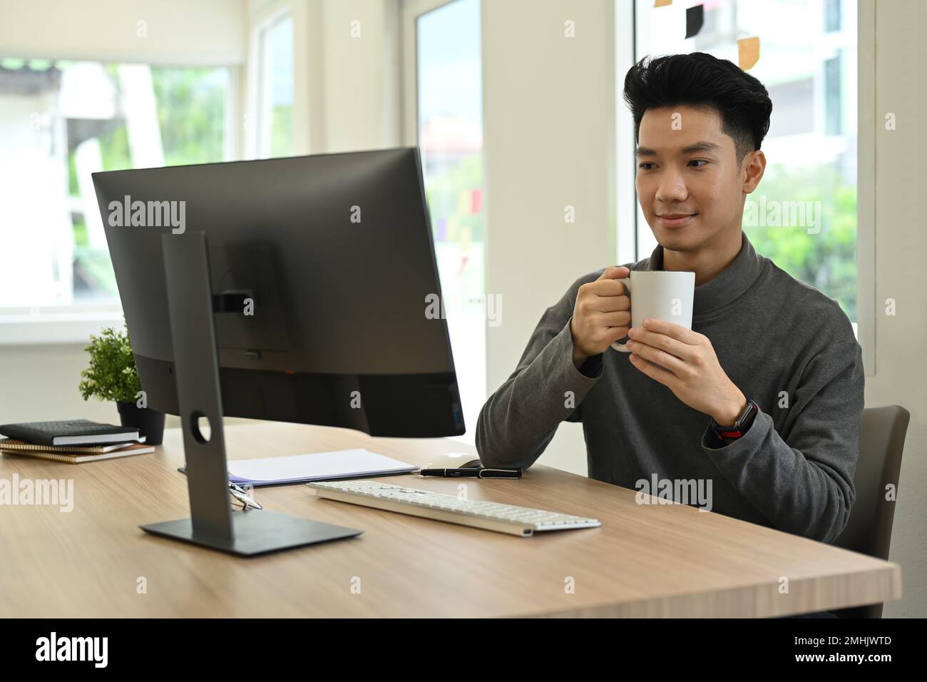 Simpatico giovane uomo asiatico freelancer bere caffè caldo e guardare lo schermo del computer Foto Stock