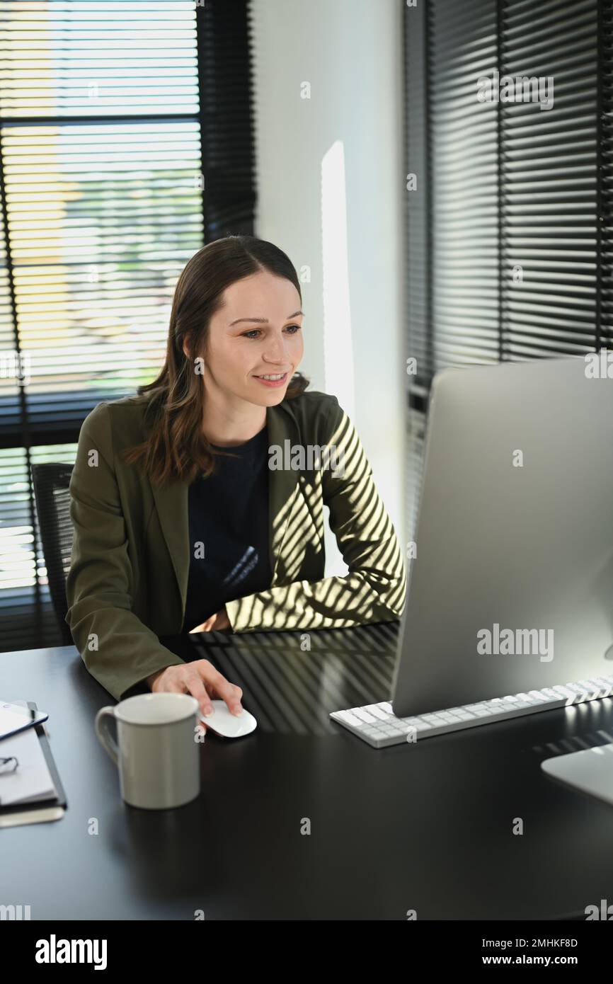 Giovane donna economista caucasica sorridente che analizza il mercato finanziario sullo schermo del computer mentre si siede alla sua postazione di lavoro Foto Stock