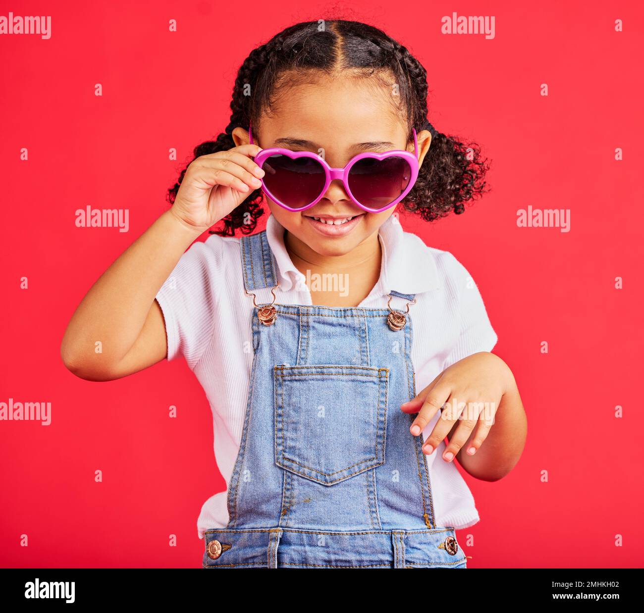 Bambina, occhiali da sole moda e cuore su sfondo rosso isolato in tendenza bambini, stile di vacanza o fresco estate. Sorridere, felice e capretto con Foto Stock