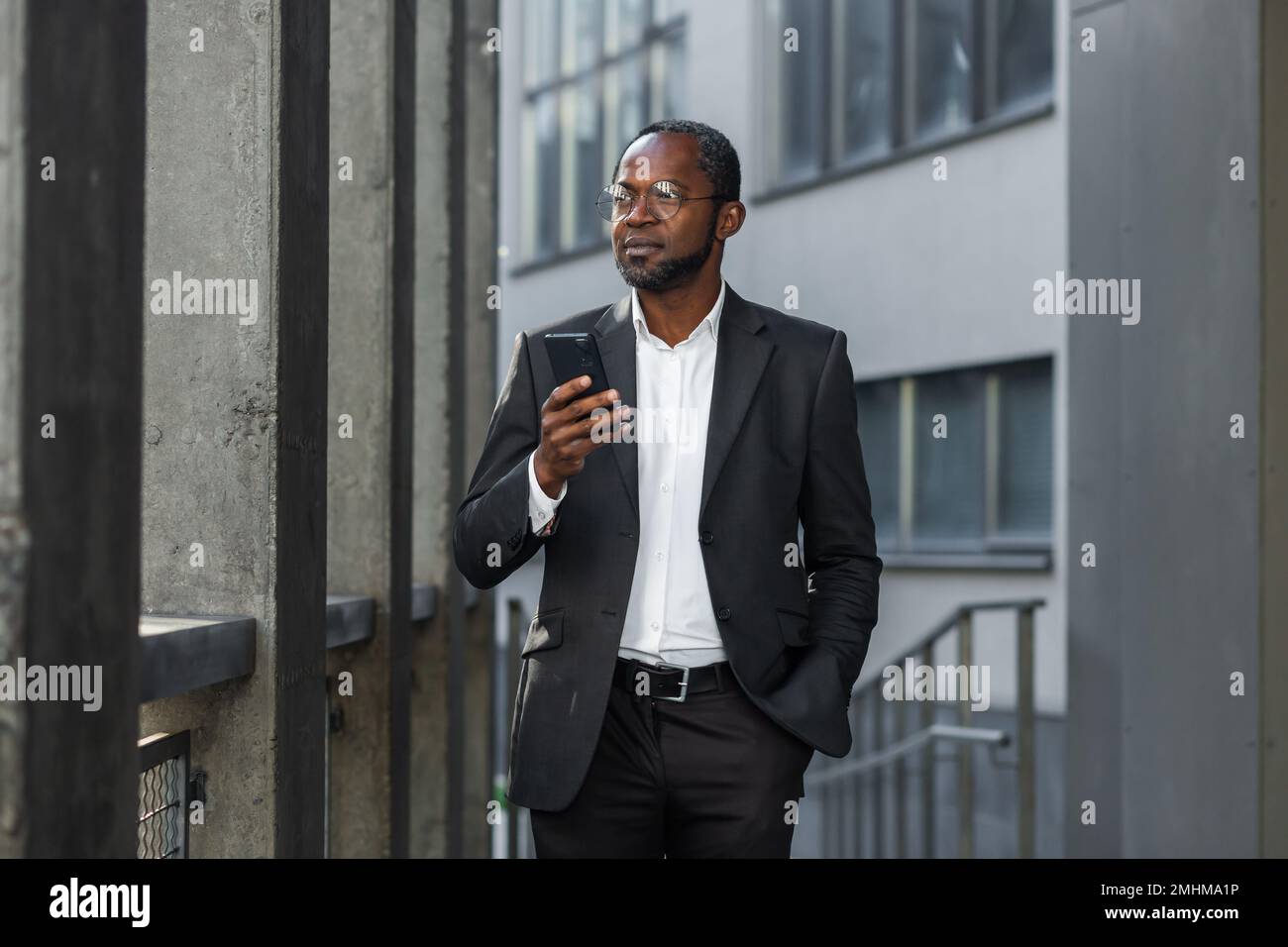 Serio e premuroso uomo d'affari che legge le notizie in linea sul boss afroamericano del telefono in vestito fuori dell'edificio dell'ufficio che osserva la concentrazione dello schermo dello smartphone, camminando vicino all'edificio in background. Foto Stock