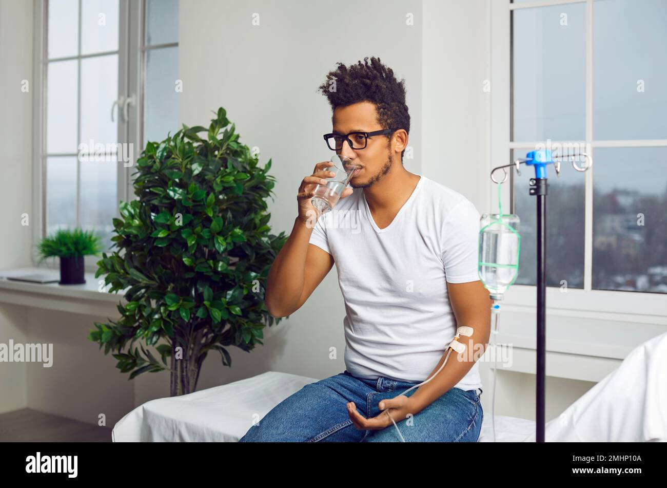 Giovane uomo indiano seduto in reparto medico sotto contagocce e bere un'acqua. Foto Stock