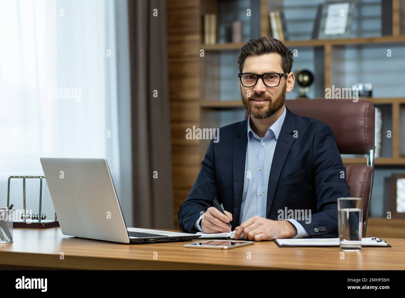 Ritratto di un uomo sorridente di successo in ufficio, uomo d'affari maturo guardando la fotocamera allegramente, capo senior in occhiali e barba lavorando in ufficio con computer portatile e documenti, firma del contratto. Foto Stock