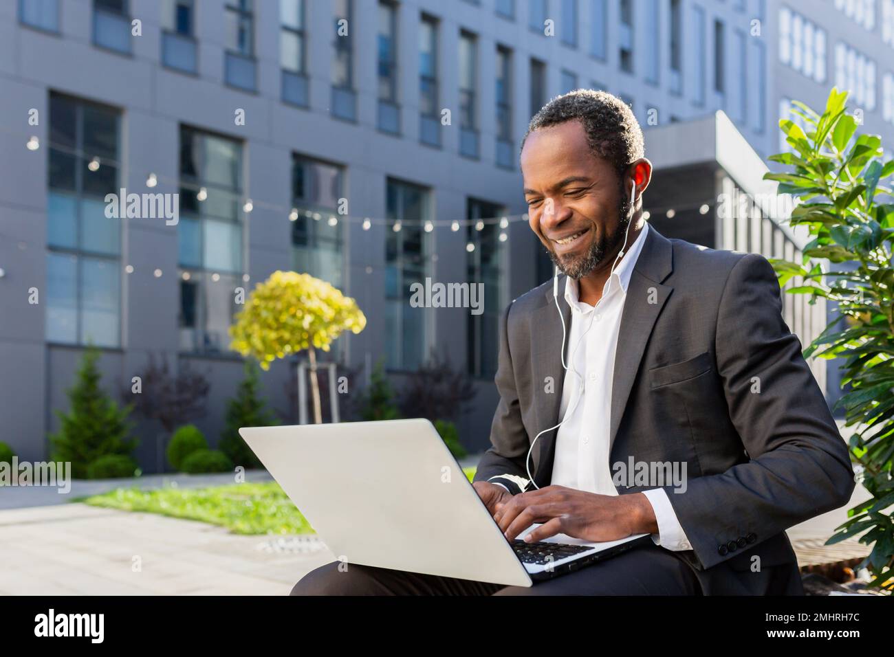 Uomo d'affari, freelancer afroamericano seduto vicino al centro dell'ufficio, che lavora online su un computer portatile e indossa le cuffie. Digita sulla tastiera, comunica tramite videochiamata, sorride. Foto Stock