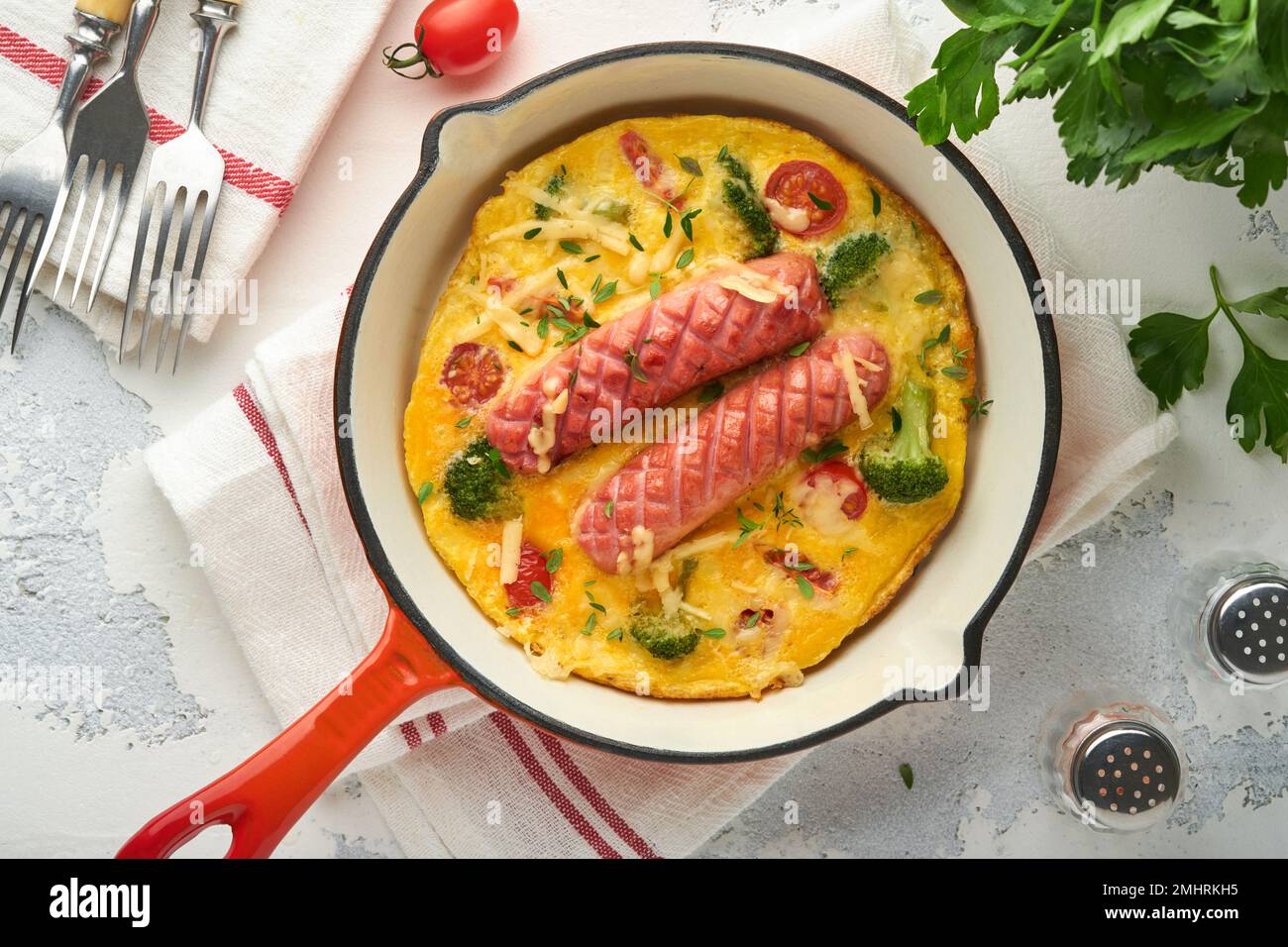 Omelette o frittata con due salsicce, formaggio fuso, pomodori ciliegini, broccoli e prezzemolo su padella rossa o teglia di ferro su fondo bianco di cemento vecchio Foto Stock