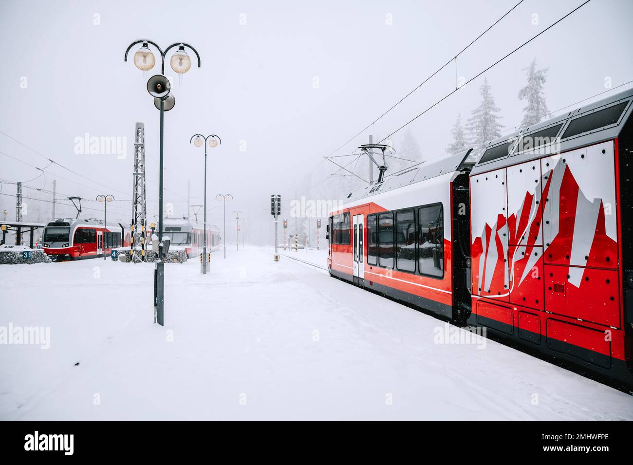 Un treno cremisi si trova a riposo su una stazione innevata nel cuore dei maestosi alti Tatra. Il motore rosso contrasta con l'inverno bianco Foto Stock