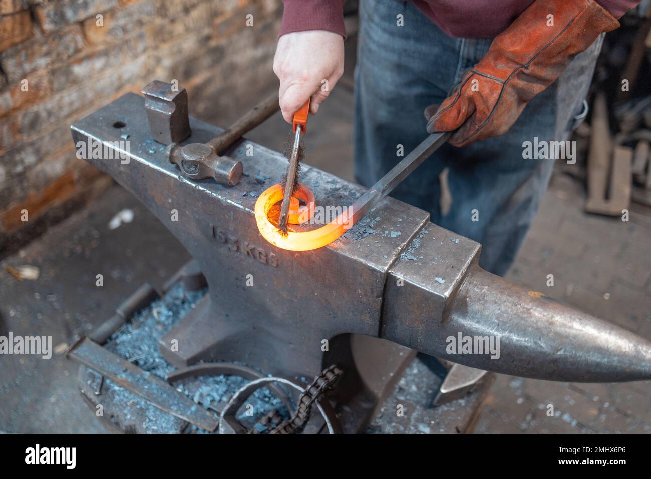 Un uomo con un guanto che tiene un martello di ferro caldo su una vecchia incudine Foto Stock