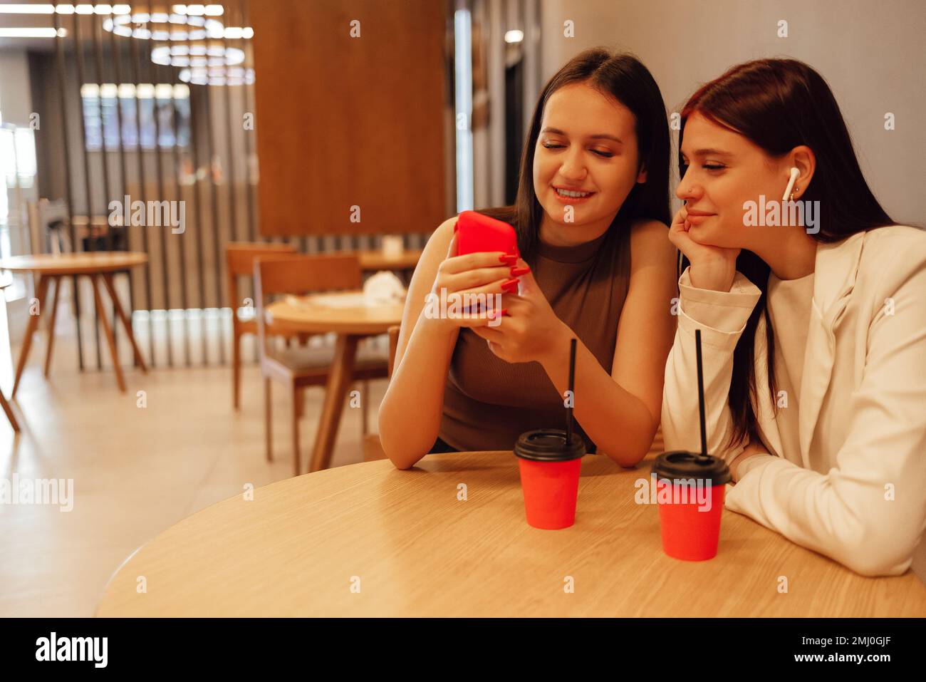 Due ragazze sono sedute in un cafè con interni moderni. Donne sorridenti brunette longhaired che guardano lo smartphone al ristorante. Le amiche bevono Foto Stock