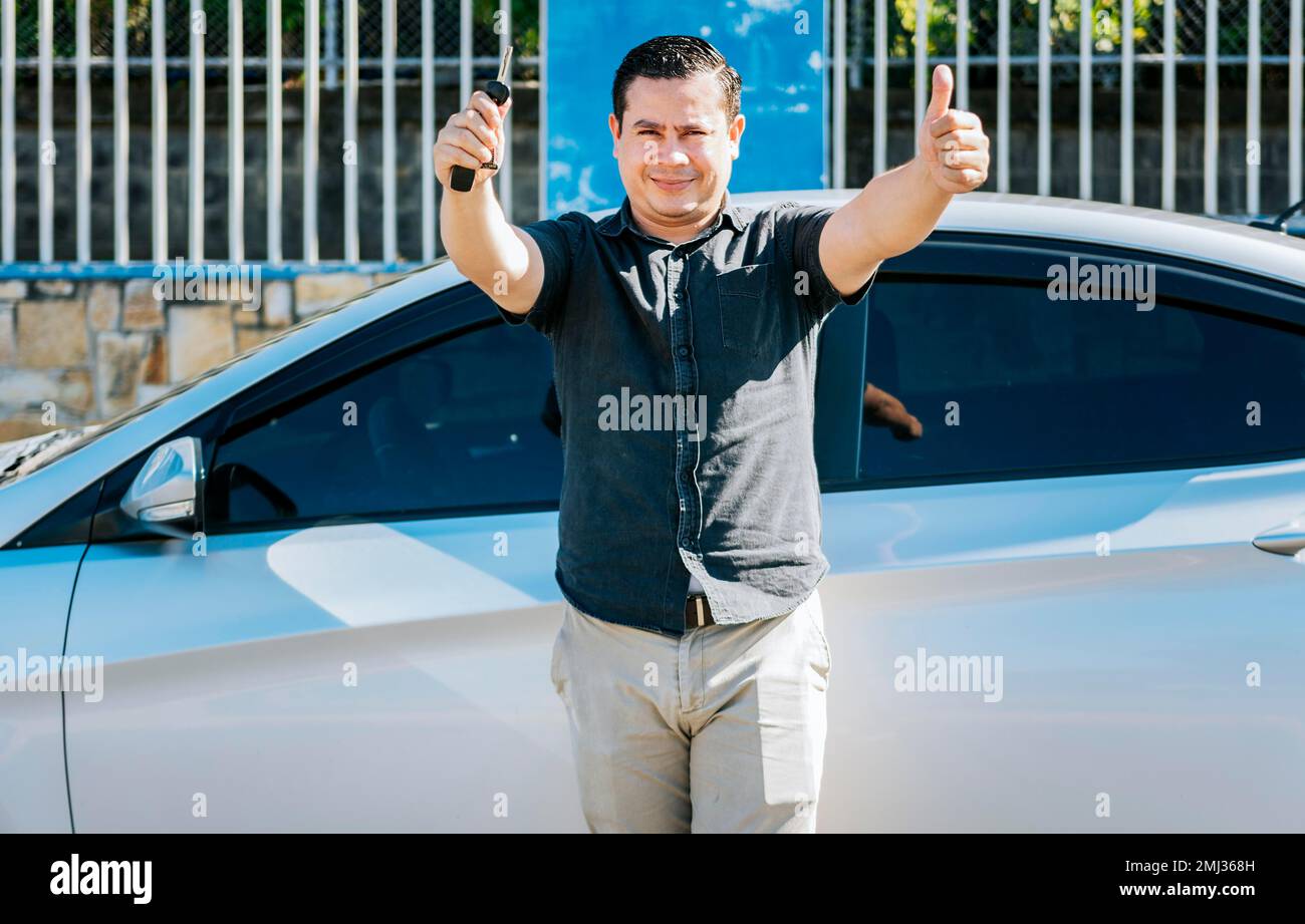 Conducente maschio in piedi accanto alla vettura mostrando le chiavi con il pollice in alto. Sorridente proprietario con la nuova auto in piedi mostrando le chiavi, conducente appoggiato sulla vettura Foto Stock