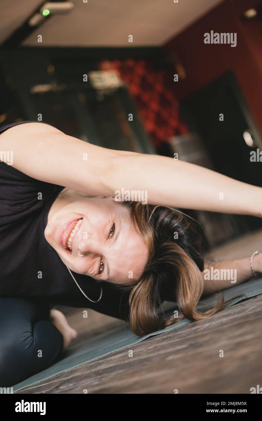 Foto ravvicinata di una nuova insegnante di yoga femminile che ha una vestibilità ridente mentre fa un tratto di corpo laterale seduto indossando una t-shirt nera all'interno Foto Stock
