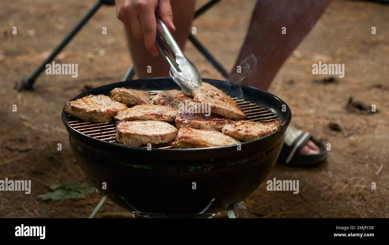 L'uomo non riconosciuto sta cucinando i pali del barbecue sulla griglia del carbone nella foresta. Preparazione del cibo da campeggio. Picchetti freschi e succosi grigliati sul barbecue. Foto Stock