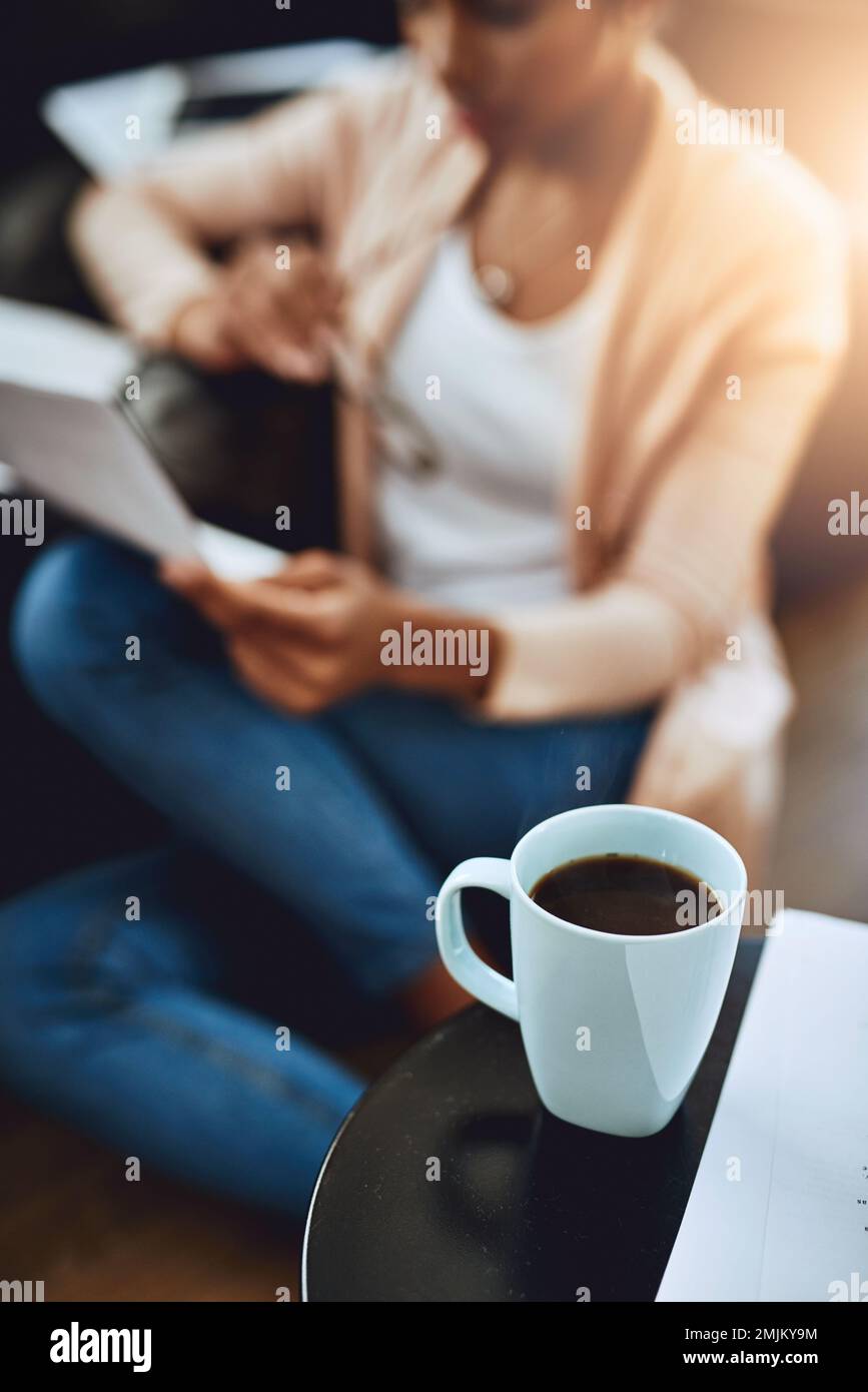 Home è dove si trova. una tazza di caffè su un tavolo con una donna che legge un documento sullo sfondo a casa. Foto Stock