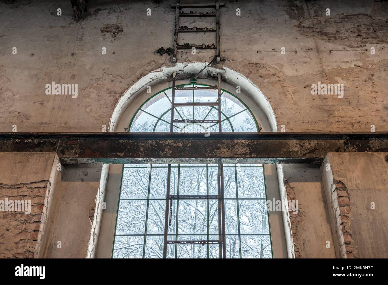Alta finestra porticato in una vecchia fabbrica. Trave a i in acciaio nella parete in mattoni e scala in metallo fissata alla parete. Paesaggio invernale fuori dalla finestra. Arrugginito, Foto Stock