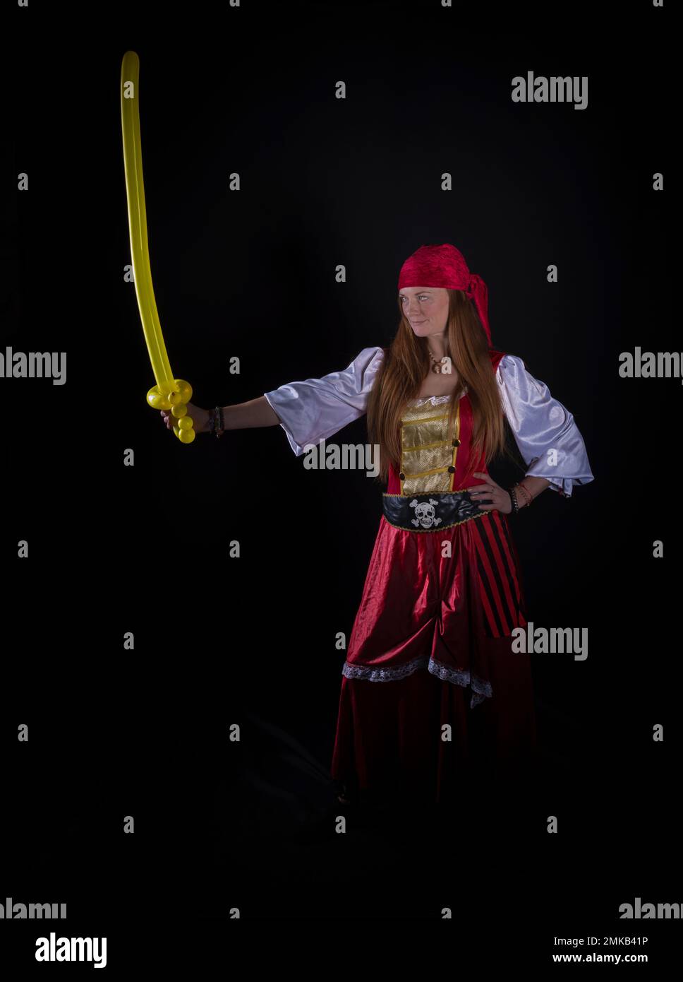 donna con capelli lunghi vestita da pirata in tuta rossa e con un pallone giallo a forma di spada nella fotografia a mano di eventi di festa Foto Stock