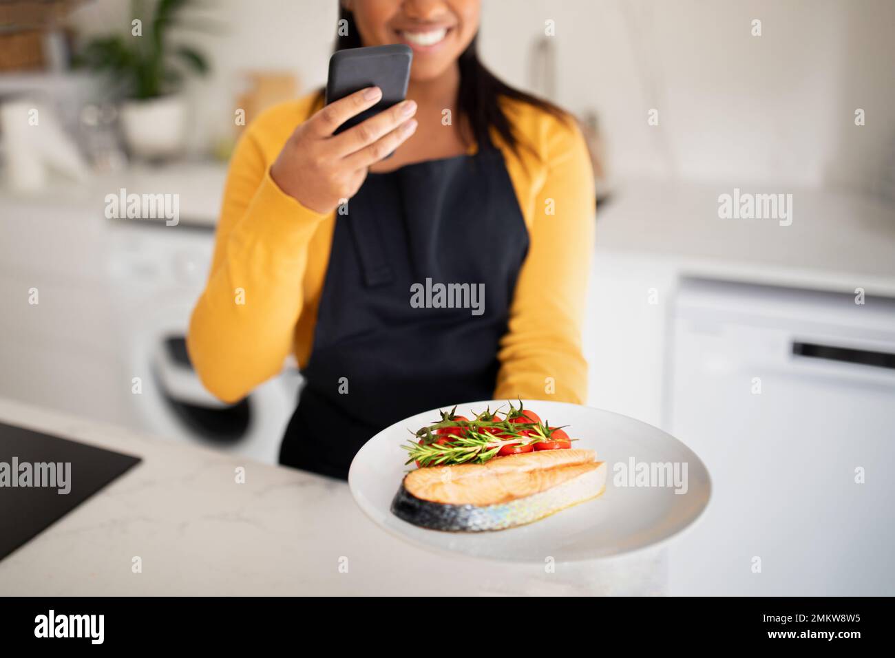 Allegra signora afroamericana millenaria in grembiule scattando una foto del piatto di pesce su smartphone Foto Stock
