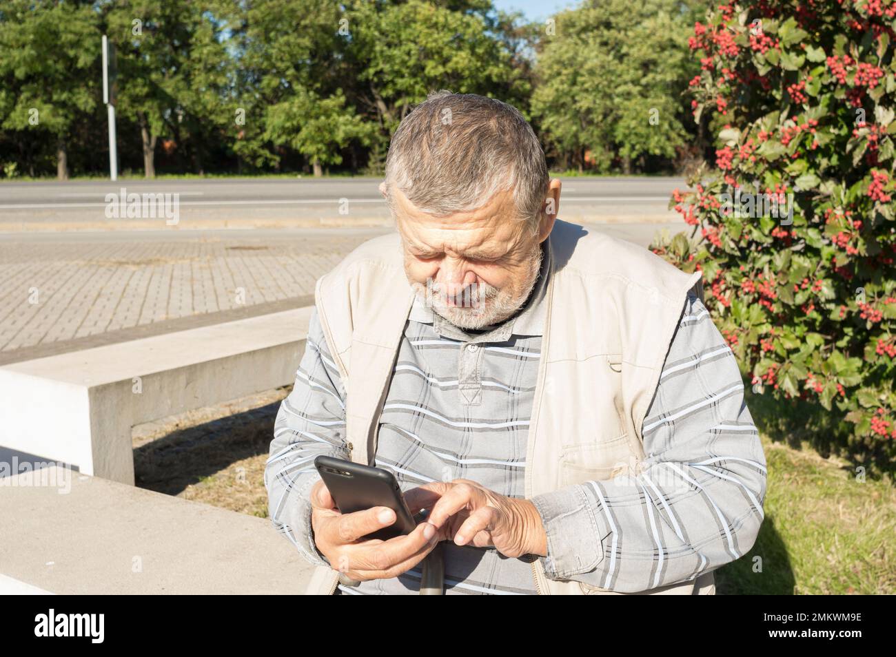 Uomo caucasico anziano seduto su panchina e utilizzando cellulare al giorno autunnale soleggiato in Ucraina Foto Stock