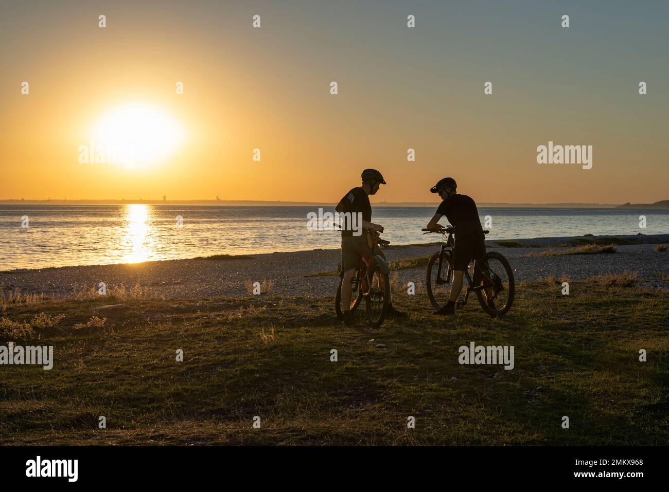 Due ciclisti retroilluminati che parlano al tramonto sulla spiaggia di Sletterhage, Djursland, Jutland, Danimarca Foto Stock