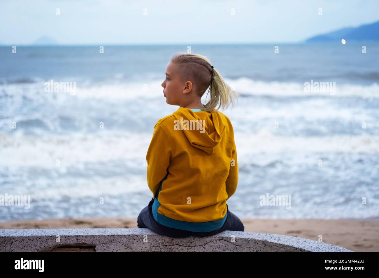 Solitario adolescente che guarda al mare seduto sul lungomare. Vista posteriore dell'adolescente contro il seascape. Foto Stock